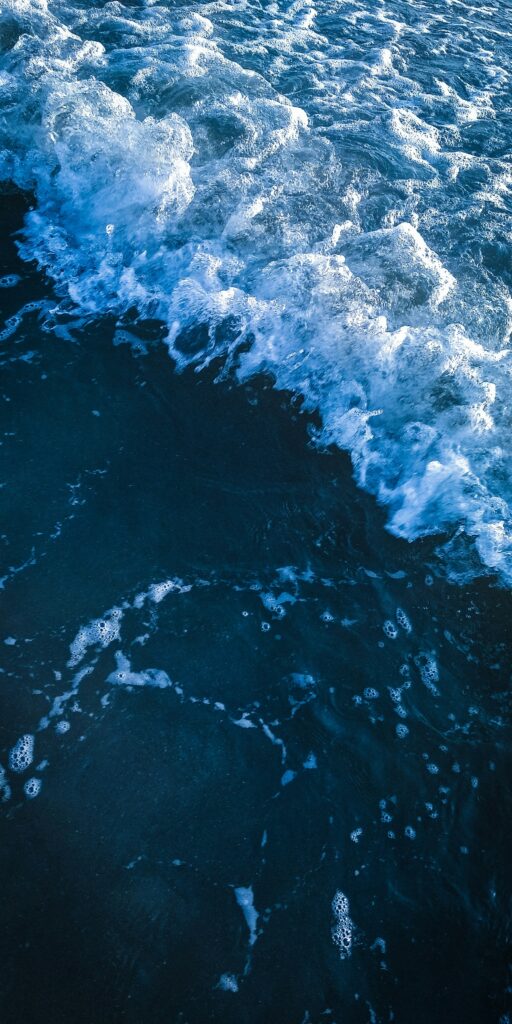 Gros plan sur des vagues océaniques dont l'écume blanche déferle sur une eau bleu foncé, créant un contraste texturé et dynamique entre l'écume des vagues et les profondeurs de la mer en contrebas.