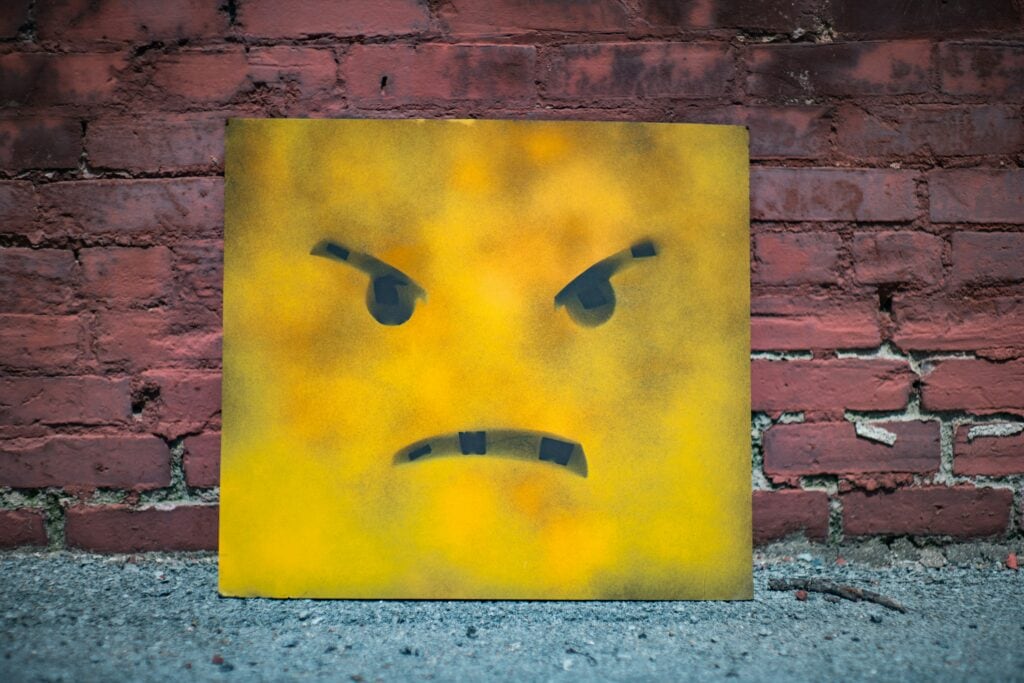 Une toile carrée jaune, sur laquelle est peinte à la bombe une tête en colère de couleur noire, symbolisant le stress scolaire, est appuyée contre un mur de briques rouges, sur un sol en béton.