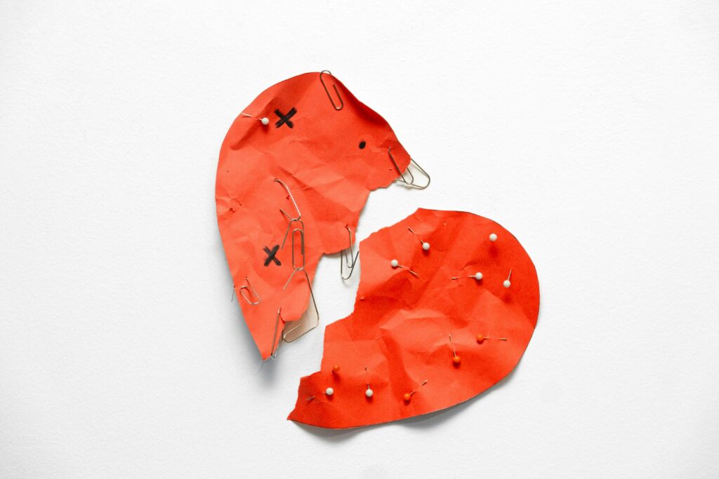 Un cœur en papier rouge, déchiré en deux et maintenu par des trombones et des épingles blanches, repose sur une surface blanche. Les morceaux du cœur sont froissés, et de petits X sont dessinés sur l'une des moitiés.