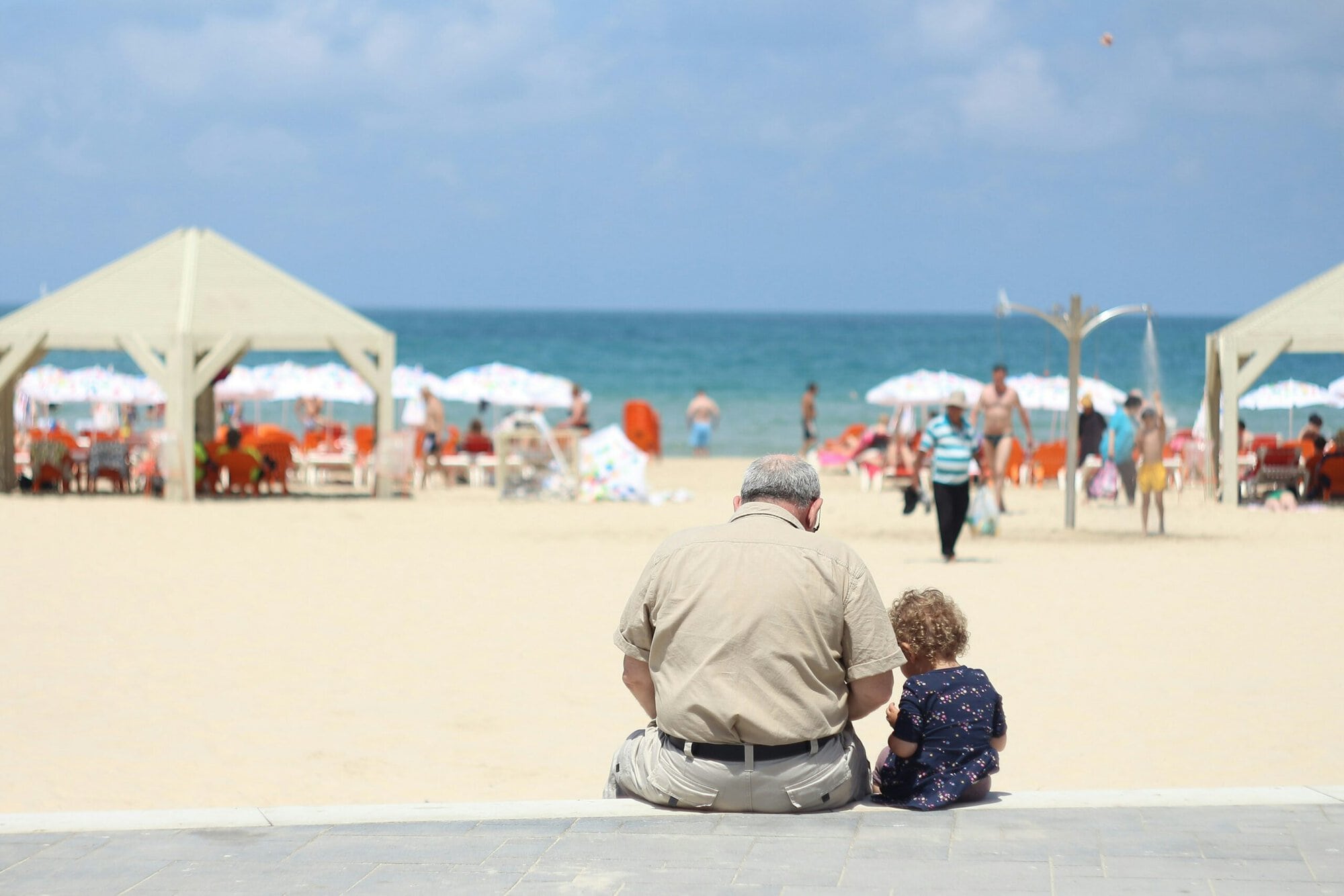 Un homme d'un certain âge et un enfant sont assis sur un rebord pavé face à une plage de sable, avec en arrière-plan des parasols, des gens et l'océan, sous un ciel partiellement nuageux.