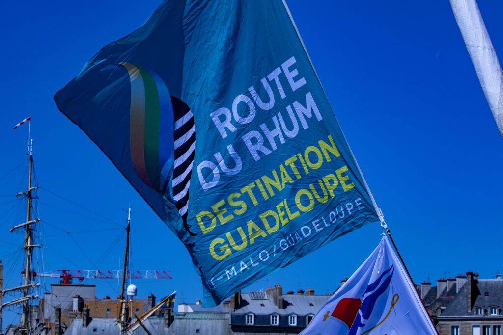 Un grand drapeau bleu de la Route du Rhum Destination Guadeloupe flotte dans un ciel bleu limpide, au-dessus des immeubles et des mâts des bateaux.