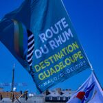 Un grand drapeau bleu de la Route du Rhum Destination Guadeloupe flotte dans un ciel bleu limpide, au-dessus des immeubles et des mâts des bateaux.