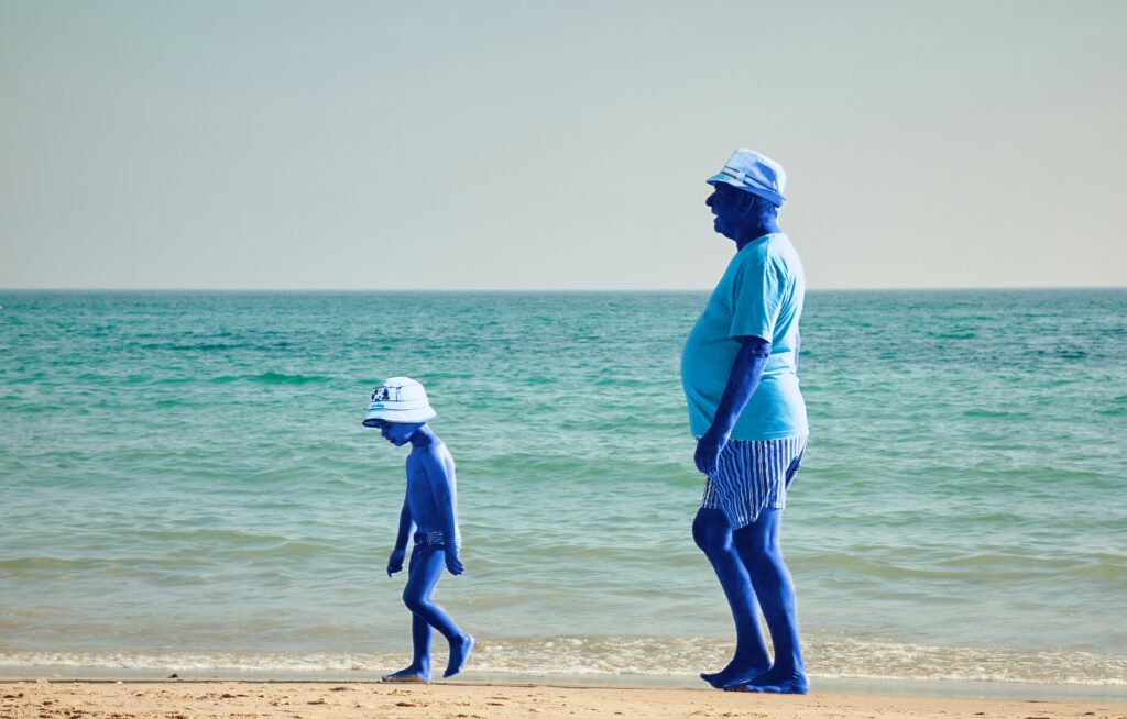 Un homme d'un certain âge et un enfant, tous deux à la peau bleue, marchent le long d'une plage de sable près de l'eau, coiffés de chapeaux et vêtus de tenues d'été. On aperçoit l'océan et le ciel en arrière-plan.