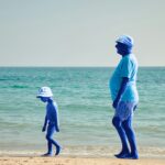 Un homme d'un certain âge et un enfant, tous deux à la peau bleue, marchent le long d'une plage de sable près de l'eau, coiffés de chapeaux et vêtus de tenues d'été. On aperçoit l'océan et le ciel en arrière-plan.