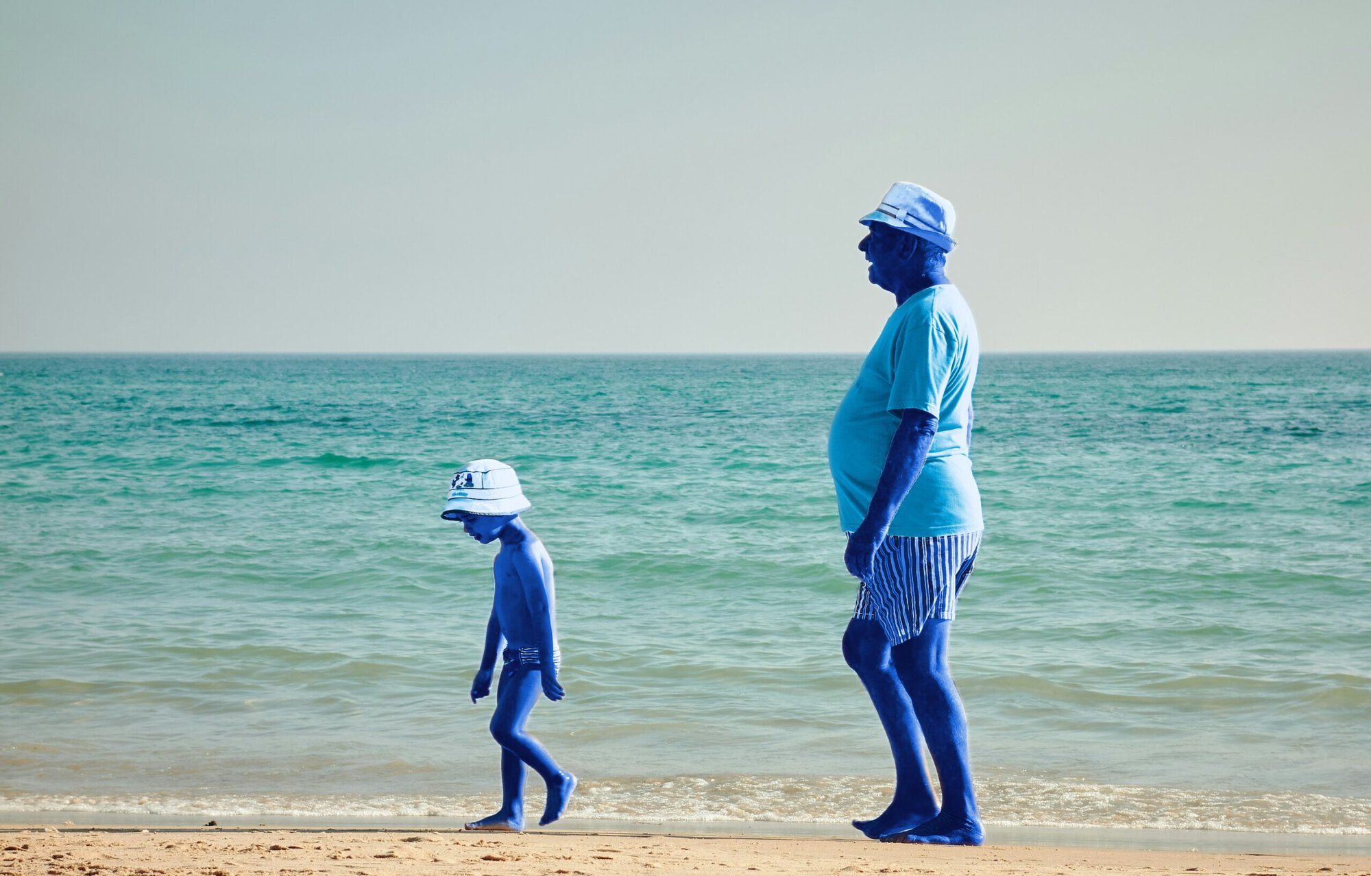 Un homme d'un certain âge et un enfant, tous deux à la peau bleue, marchent le long d'une plage de sable près de l'eau, coiffés de chapeaux et vêtus de tenues d'été. On aperçoit l'océan et le ciel en arrière-plan.