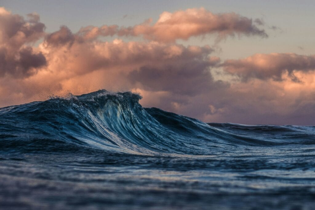 Gros plan sur une vague bleue déferlant sous un ciel spectaculaire parsemé de nuages roses au coucher du soleil.
