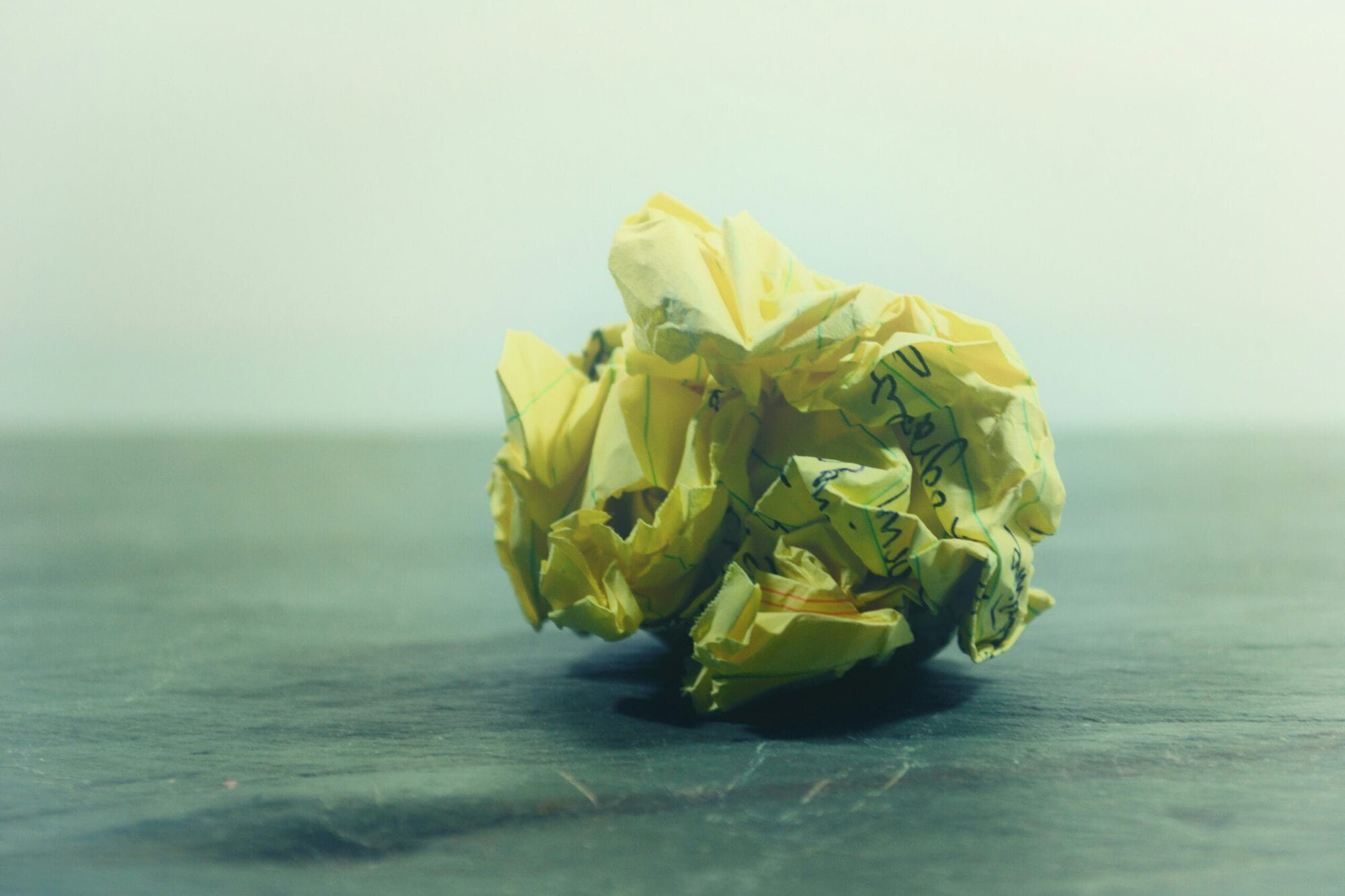 Un morceau de papier jaune froissé, sur lequel figure un texte écrit à la main, repose sur une surface sombre et lisse, devant un arrière-plan clair et flou.