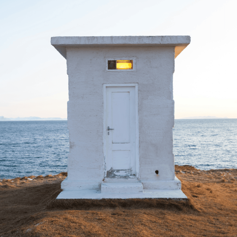 Une petite construction en béton blanc, dont la porte est fermée et au-dessus de laquelle brille une lampe, se dresse sur de l'herbe sèche près d'un plan d'eau tranquille, encadrée au loin par des montagnes enneigées sous un ciel pâle.