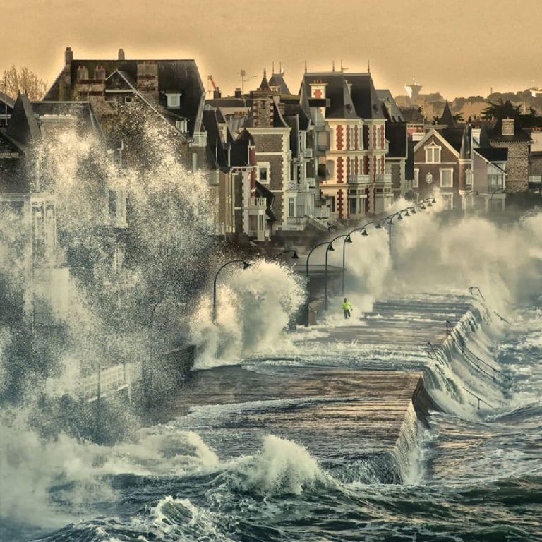 De grosses vagues viennent s'écraser contre une promenade en bord de mer, projetant des gerbes d'eau bien au-dessus des maisons du littoral sous un ciel nuageux ; le temps orageux a inondé la rue, et la mer déchaînée domine la scène.