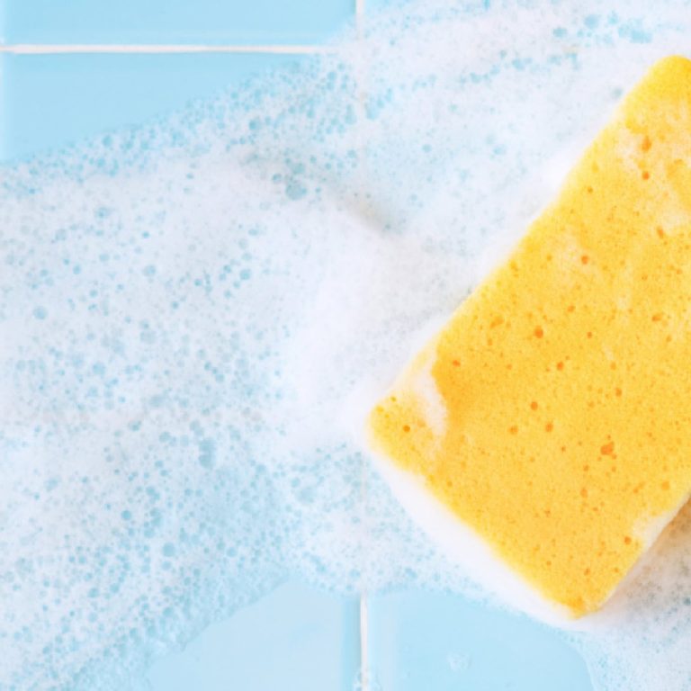 Une éponge jaune recouverte de mousse de savon repose sur une surface carrelée bleu clair, entourée de mousse, ce qui suggère qu'un nettoyage est en cours.