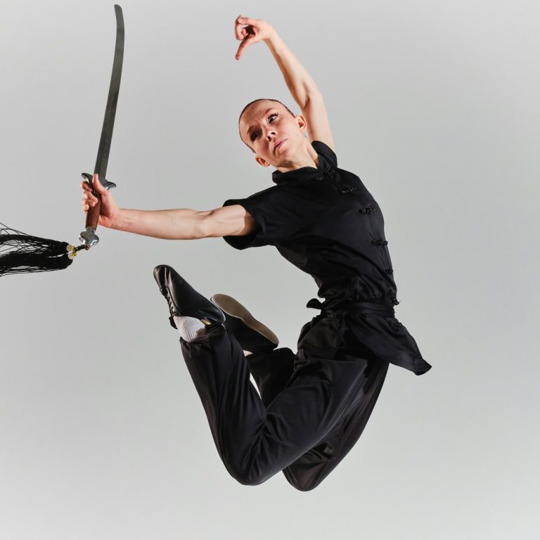 Une personne vêtue d'une tenue noire d'arts martiaux effectue un grand saut, une épée à la main, les jambes fléchies et les bras tendus, sur un fond clair et uni, mettant en valeur le dynamisme du mouvement et la concentration.