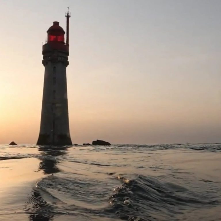 Au coucher du soleil, un grand phare au sommet rouge se dresse au milieu de la mer. Le soleil est bas à l'horizon, projetant une lueur chaude sur l'eau calme et créant de légères ondulations au premier plan.