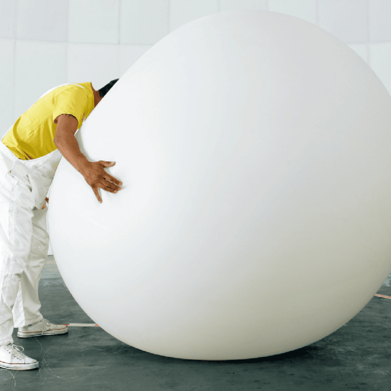 Une personne vêtue d'une chemise jaune et d'un pantalon blanc se penche et enlace un grand objet blanc, lisse et en forme d'œuf, dans un espace intérieur moderne et presque vide.