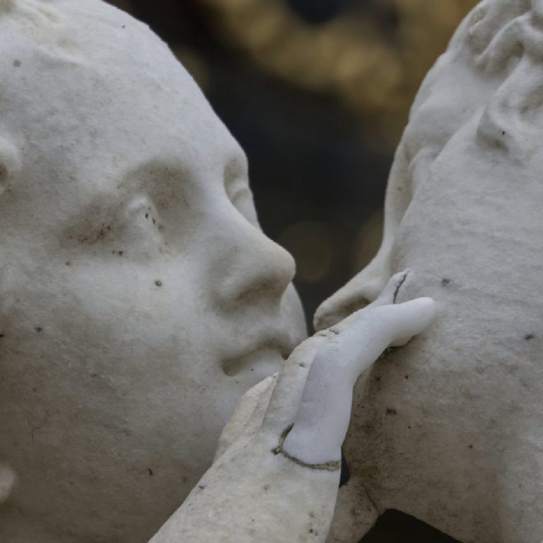 Gros plan sur une statue de marbre représentant deux figures angéliques, dont l'une effleure doucement le visage de l'autre, aux traits doux et aux détails finement sculptés qui soulignent leur beauté angélique.