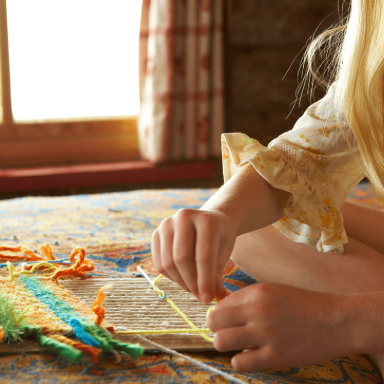 Une jeune fille est assise en tailleur sur le sol, tissant des fils aux couleurs vives sur un petit métier à tisser, près d'une fenêtre baignée de soleil aux rideaux à motifs.