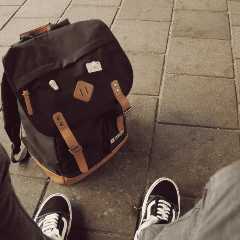 Une personne vêtue d'un pantalon gris et de baskets noires est assise sur un trottoir carrelé, face à un sac à dos noir robuste muni de sangles marron, posé à la verticale sur le sol.