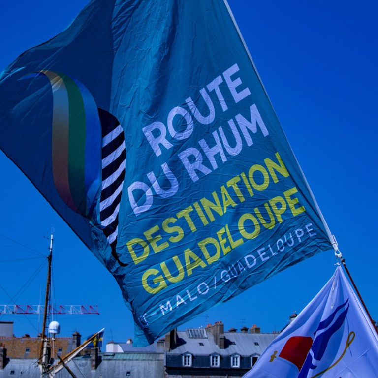 Un grand drapeau bleu de la Route du Rhum Destination Guadeloupe flotte dans un ciel bleu limpide, au-dessus des immeubles et des mâts des bateaux.