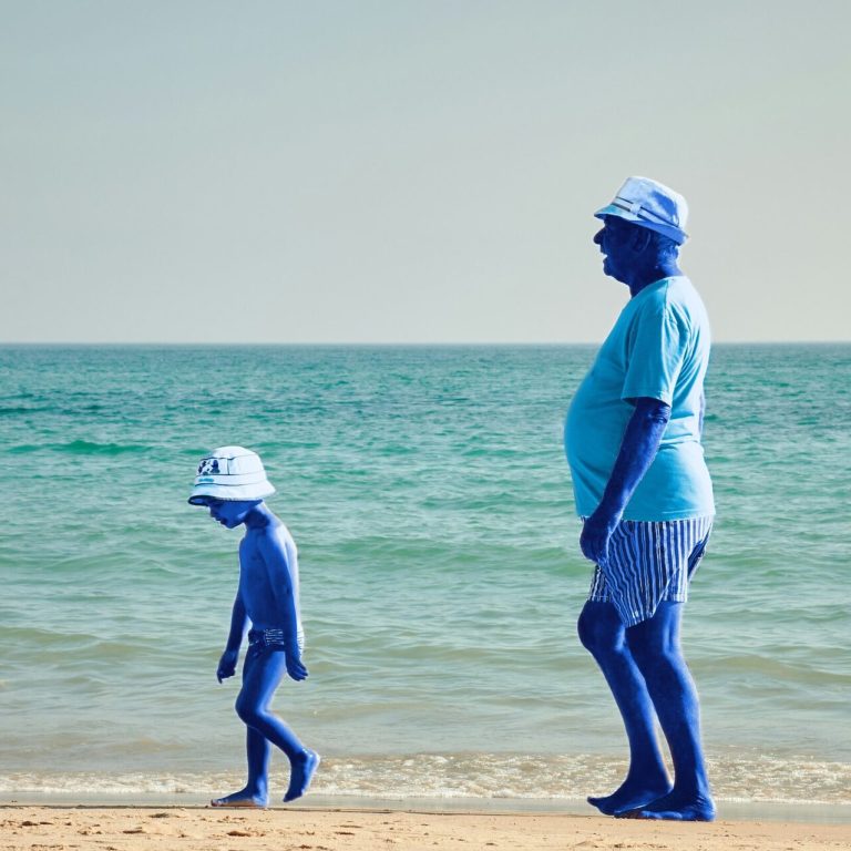 Un homme d'un certain âge et un enfant, tous deux à la peau bleue, marchent le long d'une plage de sable près de l'eau, coiffés de chapeaux et vêtus de tenues d'été. On aperçoit l'océan et le ciel en arrière-plan.